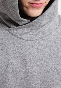 Man draagt een grijze trui met capuchon van een zacht, gestructureerd materiaal, waarbij alleen het onderste deel van het gezicht en de nek zichtbaar zijn.