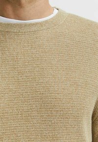 Maglione a maglia color beige con un motivo testurizzato simile a una cialda. Collo rotondo sopra una camicia bianca a strati, che mette in mostra la morbidezza del tessuto.