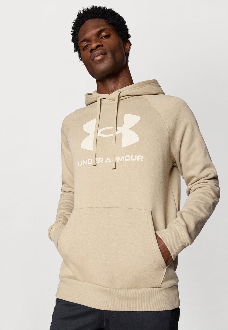Uomo che indossa un felpa beige Under Armour con tasca frontale e cordini, in piedi con le mani in tasca contro uno sfondo semplice.