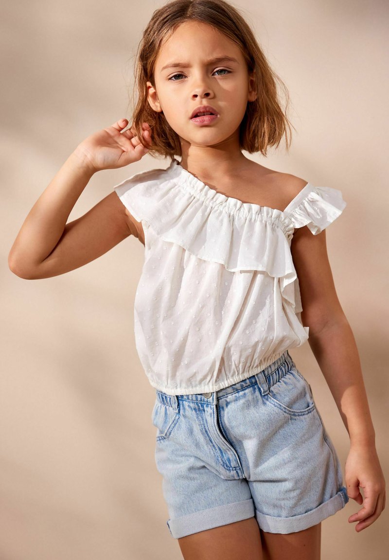 Weißes, rüschengeschnittenes Top mit einem Schulterträger und subtiler Punktstruktur, kombiniert mit hellblauen Denim-Shorts mit umgeschlagenem Saum und elastischem Bund.