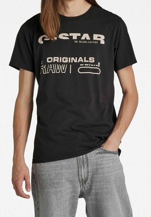 Osoba ubrana w czarną koszulkę z logo G-Star RAW Originals i jasnoszare dżinsy, z jedną ręką w kieszeni, na jednolitym tle.