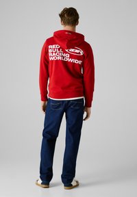 Rote Kapuzenjacke mit großem weißen Text "RED BULL RACING WORLDWIDE" auf dem Rücken, kombiniert mit blauen Jeans und lässigen Schuhen.