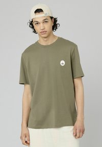 T-shirt en coton vert olive avec une coupe décontractée, des manches courtes, un col rond et un petit patch circulaire représentant un voilier.
