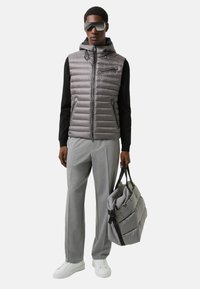 Graue Steppweste mit Kapuze, mit Reißverschlusstaschen, kombiniert mit grauen Hosen und weißen Sneakers. Hält eine passende graue Tasche mit Trägern.