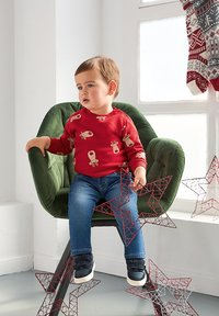 Camicetta rossa a maniche lunghe con stampa di renne, jeans blu, seduto su una sedia di tessuto verde, tiene in mano stelle di filo rosse e bianche.