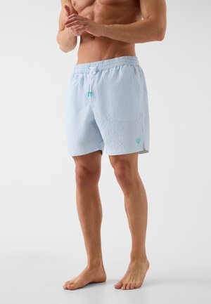 Homme musclé portant un short de bain bleu clair texturé avec un cordon blanc, debout pieds nus sur un fond neutre.