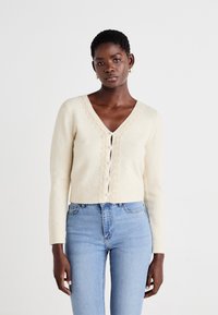 Beige strikket cardigan med V-hals, blonder langs halslinjen og fronten, og perlemorsknapper; brukt over lyseblå høyt liv jeans.