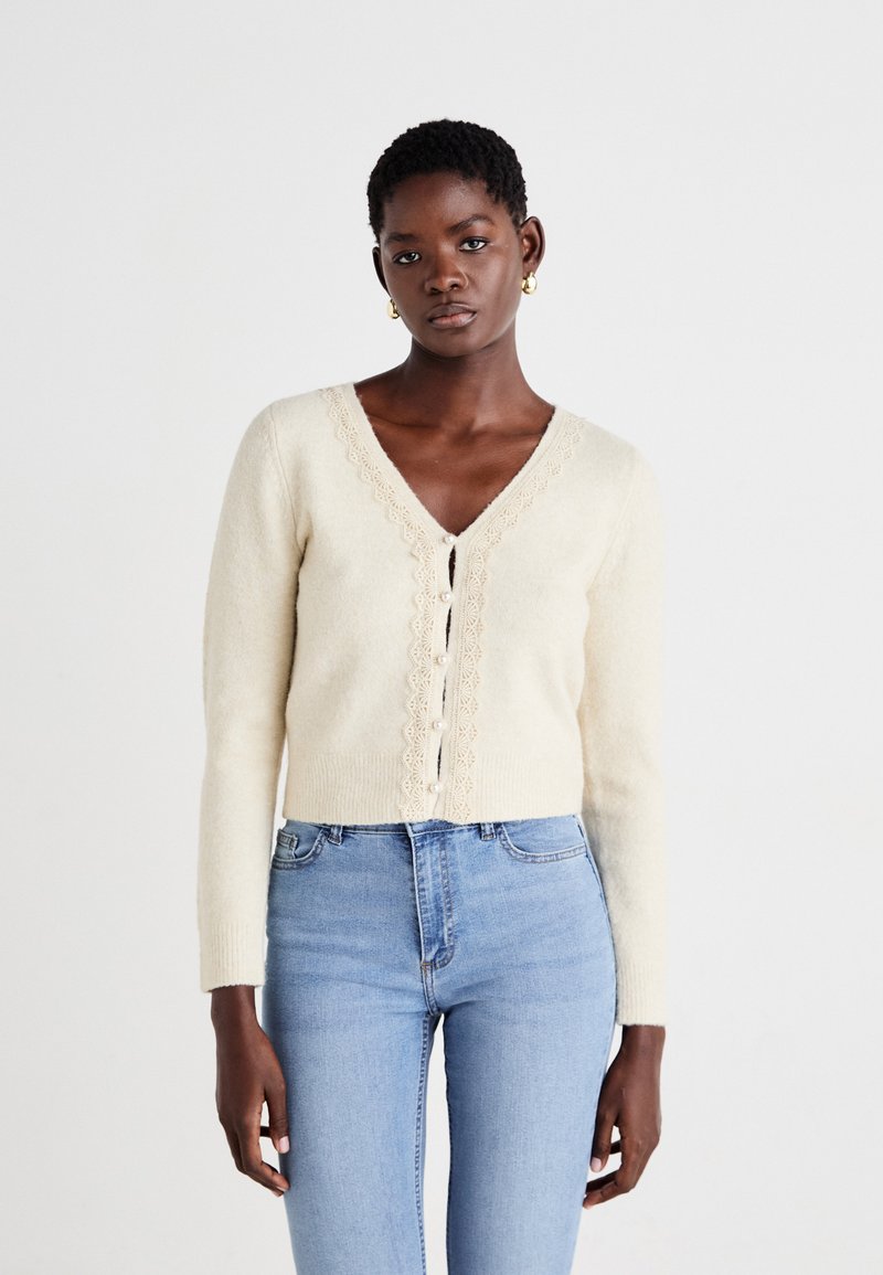 Beige strikket cardigan med V-hals, blonder langs halslinjen og fronten, og perlemorsknapper; brukt over lyseblå høyt liv jeans.