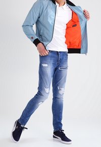 Svetle modrá bomber bunda s oranžovou podšívkou, nošená přes bílou polo košili; obnošené modré džíny; černé tenisky.