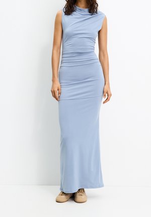 Femme portant une robe maxi bleu clair sans manches, froncée, associée à des baskets beige, debout sur un fond blanc uni.