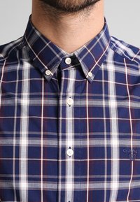 Koszula na guziki w granatowym kolorze z wzorem w kratę w czerwonym i białym kolorze. Posiada kołnierz typu spread i małe białe guziki. Logo marki na piersi.