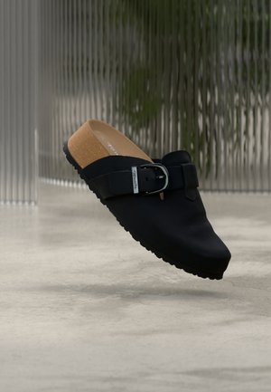 Sort slip-on træsko med korksål, justerbar spænde, og tekstureret sål, svævende over en beton gulv.