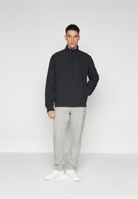 Chaqueta negra con cremallera y cuello alto, combinada con pantalones de chándal gris claro y zapatillas blancas. Diseño simple, sin patrones ni logotipos visibles.