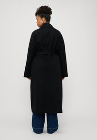 Schwarzer langer Mantel mit tailliertem Gürtel, einreihiger Knopfleiste, weiten Ärmeln und Kragen. Getragen über ausgestellt geschnittenen blauen Jeans.