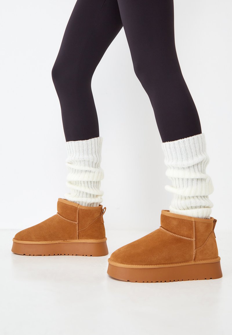 Bottines en suede beige avec une semelle en caoutchouc plate, accompagnées de jambières en laine côtelée cream. Les bottines sont dotées d'une languette à l'arrière pour un enfilage facile.