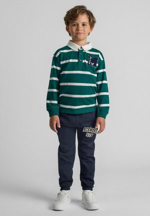 Polo a righe verdi e bianche con colletto e dettagli ricamati, abbinato a pantaloni della tuta blu navy con stampa "OREGON 1997". Sneakers bianche.