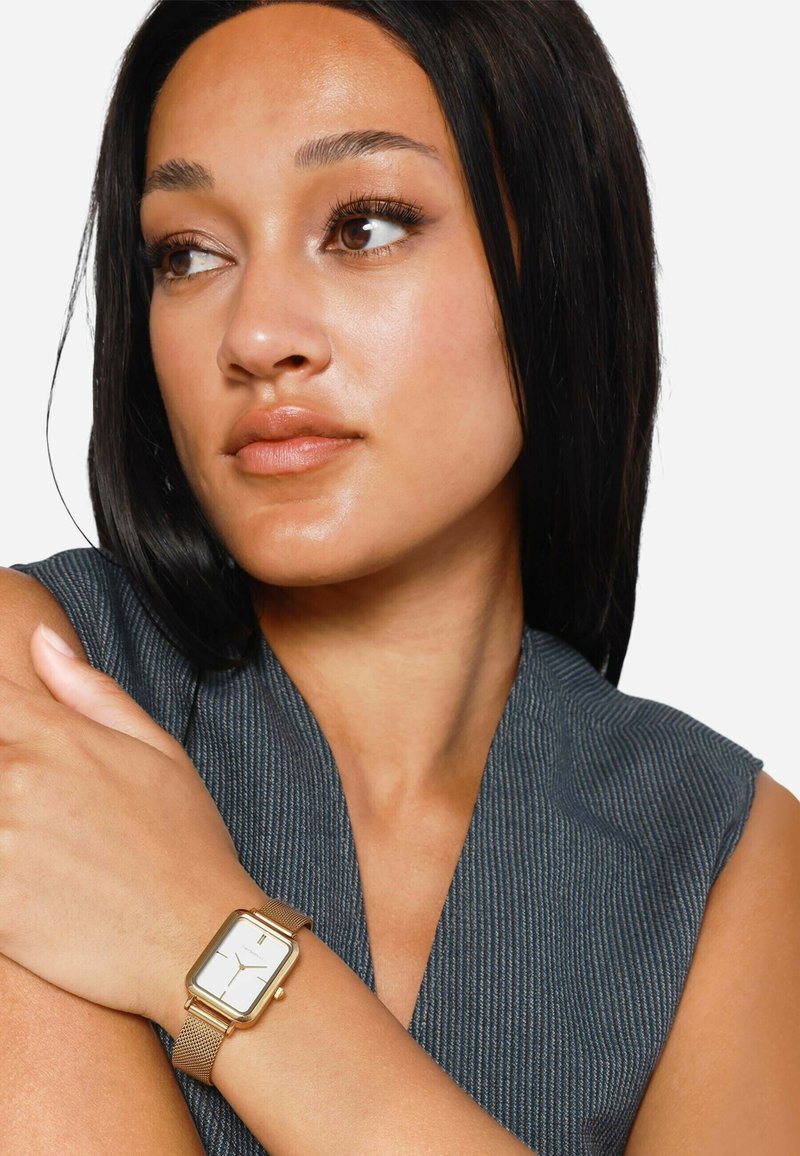 Goldfarbene rechteckige Uhr am Handgelenk, mit einem weißen Ziffernblatt und einem Mesh-Armband. Das Modell trägt ein graues, strukturiertes Oberteil, die Arme in neutraler Pose gekreuzt.