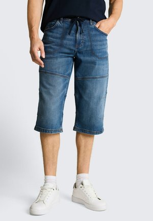 Herren blaue Denim-Kniehosen mit sichtbaren Nähten, kombiniert mit weißen Sneakers und weißen Socken, vor neutralem Hintergrund.