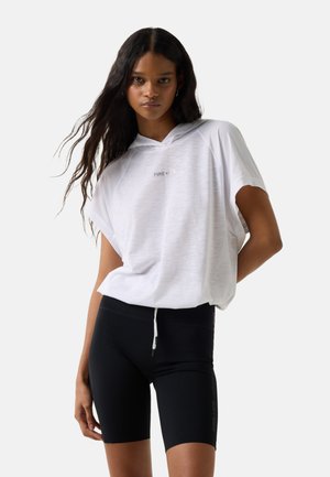 Kvinde med langt mørkt hår iført hvid kortærmet hættetrøje og sorte tætsiddende cykelshorts, stående mod en ensfarvet baggrund.