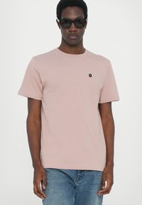Helles pinkes Baumwoll-T-Shirt mit Rundhalsausschnitt, kurzen Ärmeln und einem kleinen schwarzen sechseckigen Logo auf der linken Brust. Getragen mit blauen Jeans.