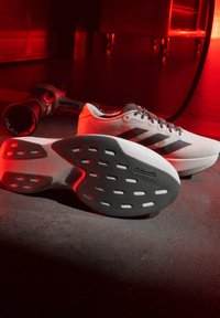 Weiße und schwarze Adidas-Laufschuhe mit Continental-Gummisohlen auf einem dunklen Boden unter roter Beleuchtung, mit einem Elektrowerkzeug im Hintergrund.