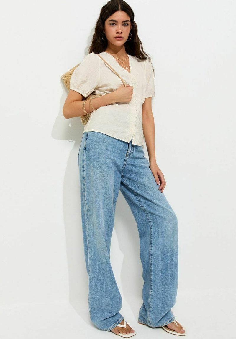 Woman with long brown hair wears a cream blouse, wide-leg blue jeans, sandals, and a straw bag, standing against a plain white background.