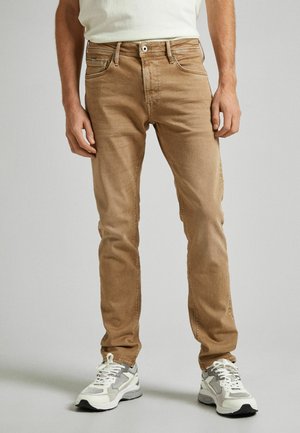 Homme portant un jean slim marron clair et des chaussures de sport blanches et grises, debout devant un fond uni gris clair.