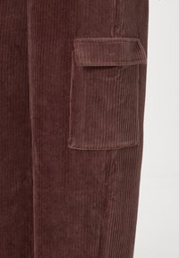 Καφέ χοντρό παντελόνι με κάθετους ραφές και μια τσέπη με πατίνα στη πλευρά. Το ύφασμα έχει απαλή υφή και ζεστό, πλούσιο χρώμα.