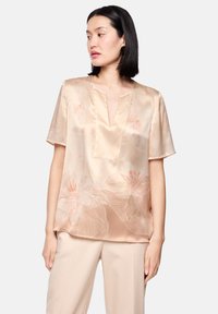 Seidenbluse in hellem Beige mit floralem Muster in zartem Rosa. Ausgestattet mit kurzen Ärmeln und einem Ausschnitt mit kleinem Schlitz sowie subtilen vertikalen Falten.