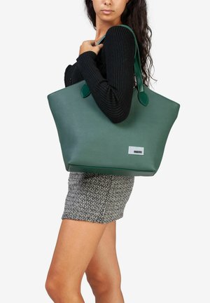 Groene nepleer tote bag met twee afgeronde handvaten, een rechthoekige vorm en een zilveren logo label aan de voorkant. Structuurstof rok zichtbaar.