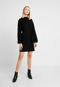 Zwart geribbeld sweaterjurk met lange mouwen, een ceintuur in de taille en contrasterende manchetten. Draag het met zwarte enkellaarsjes. Glad textuur.