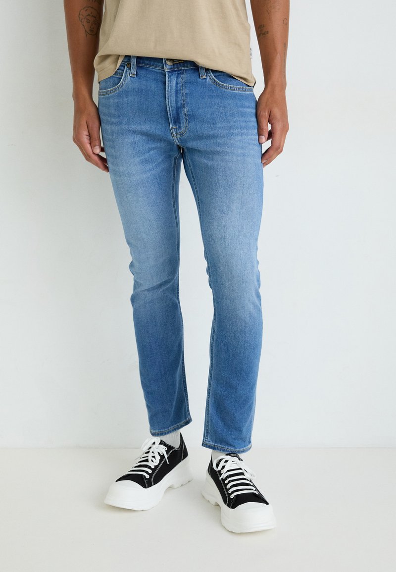 Jean en denim bleu clair à coupe slim, design à cinq poches et légère décoloration. Associé à des baskets noir et blanc.