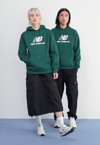 Des sweatshirts verts avec un logo blanc, un pantalon cargo noir et des baskets. Le modèle A porte une jupe, le tout sur un fond uni.