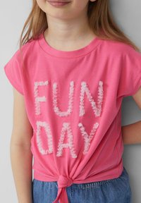 Różowa koszulka z krótkim rękawem z falbankowymi białymi literami napisanymi "FUN DAY", związana w talii, w połączeniu z dżinsowymi spodniami.