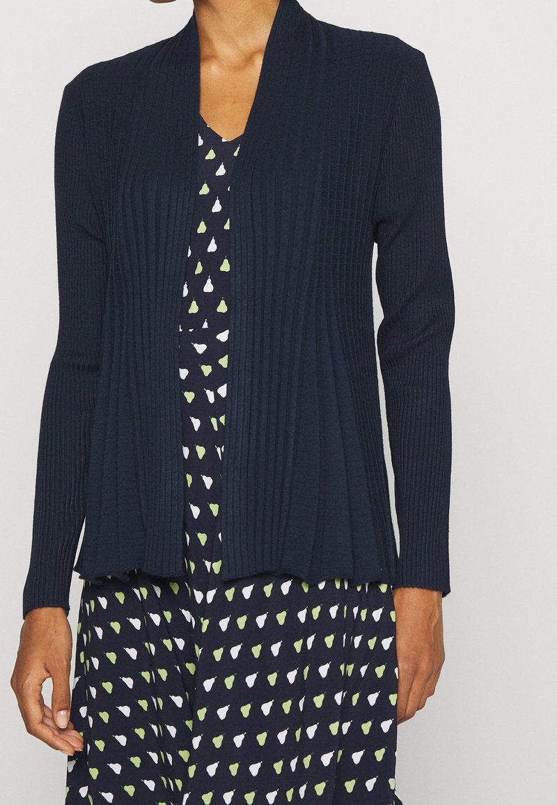 Cardigan a coste navy con apertura frontale, maniche lunghe e finitura testurizzata, abbinato a un vestito della fantasia con piccole pere.