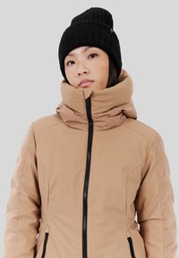 Chaqueta acolchada color beige con cuello alto, cremallera negra y bolsillos con cremallera; combinada con un gorro de punto negro acanalado. Textura suave.