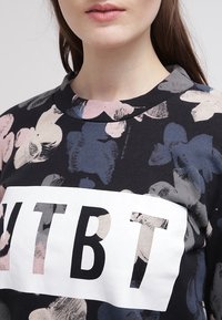 Černá bavlněná mikina s květinovým vzorem v růžové a modré barvě, s výrazným bílým grafickým nápisem "YTBT" na přední straně.