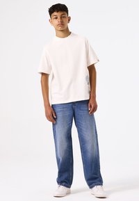 Camiseta de manga corta color beige claro con escote redondo y un gráfico impreso en el dobladillo. Combinada con unos vaqueros azules desgastados de corte holgado.
