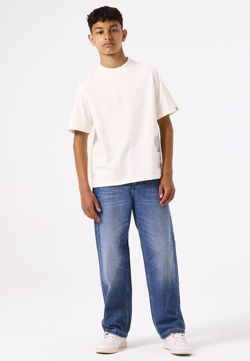Camiseta de manga corta color beige claro con escote redondo y un gráfico impreso en el dobladillo. Combinada con unos vaqueros azules desgastados de corte holgado.