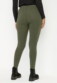 Oliivinvihreät legginsit, joissa on tiukka istuvuus, joustava vyötärö ja sileä pinta. Yhdistetty mustiin nilkkasaappaisiin, joissa on paksu pohja.