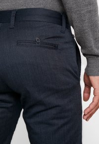 Close-up of a person wearing dark blue textured pants with a buttoned back pocket and a gray sweater sleeve visible.