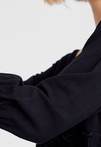 Svart texturerad blus med puffärmar, som har samlad tyg längs armen. Materialet verkar vara lätt och faller mjukt.