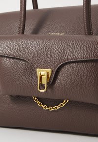 Borsa a mano in pelle marrone con superficie texture, chiusura in metallo dorato e decorativa catena dorata. Il design include manici strutturati.