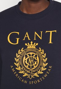 Felpa blu marina con una grande grafica gialla che presenta "GANT" e un design con un'icona circolare decorata da foglie di alloro e il testo "AMERICAN SPORTSWEAR".