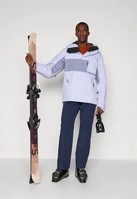 Mann iført skijakke og bukser, holder ski i den ene hånden og svarte skibriller i den andre, står mot en ensfarget bakgrunn.