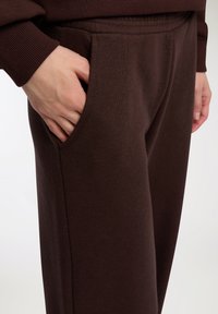 Pantaloni sportivi marroni con una texture morbida e tasca laterale. Vita elasticizzata e dettaglio a costine sul fondo. Primo piano di una mano nella tasca.