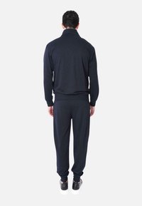 Tuta da ginnastica navy con colletto con zip, dotata di polsini e vita elasticizzati. Il tessuto appare morbido e leggermente texture. Sneakers bianche visibili.
