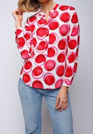 Persoon die een witte blouse met lange mouwen met grote rode cirkelpatronen draagt en lichtblauwe spijkerbroek, poseert met één hand bij het haar.