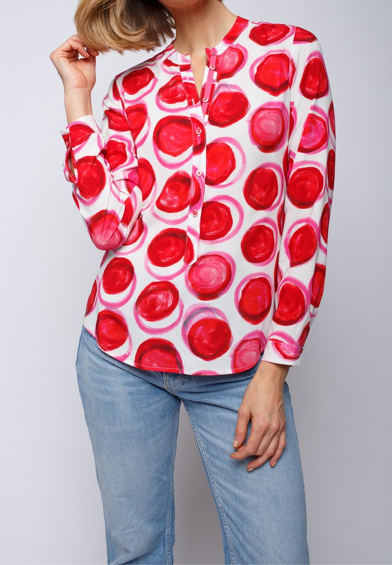 Personne portant un chemisier blanc à manches longues avec de grands motifs circulaires rouges et un jean bleu clair, posant avec une main près des cheveux.