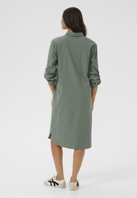 Robe chemise verte à manches longues avec col, présentant des manches retroussées, un ourlet incurvé et des fentes latérales, associée à des baskets blanches.